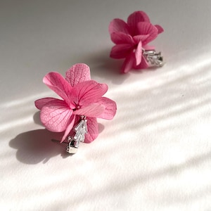 Op de afbeelding: Een paar roze bloemen oorbellen. Elke oorbel heeft een levendige roze bloem met delicate bloemblaadjes. De bloemen zijn gecombineerd met zilverkleurige, geometrisch gevormde accenten. De oorbellen staan tegen een witte achtergrond, met zachte schaduwen.