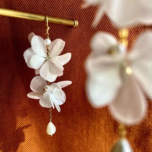 May include: Elegant white floral earrings with gold-tone hooks and chains. Each earring showcases two layered flower petals and a teardrop pearl. The earrings are set against a warm, orange-brown background.