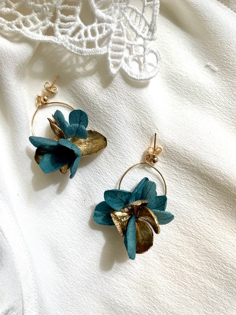 Puede incluir: Un par de pendientes de aro dorados con detalles de flores azul turquesa. Las flores est&aacute;n hechas de flores secas prensadas.