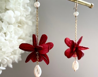 May include: A pair of dangle earrings featuring a red flower charm and a white pearl drop. The earrings are made with a gold chain and have a hook closure.