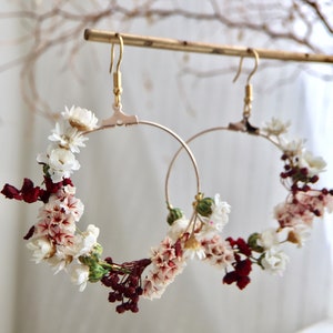 Peut inclure: Une paire de boucles d'oreilles créoles dorées décorées de fleurs séchées blanches et bordeaux. Les boucles d'oreilles sont suspendues à une barre dorée.
