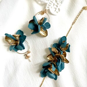 Puede incluir: Un conjunto de pendientes de flores doradas y azul turquesa y una pulsera a juego. Los pendientes son de aro con un racimo de flores sujetas. La pulsera tiene una cadena con un racimo de flores sujetas.