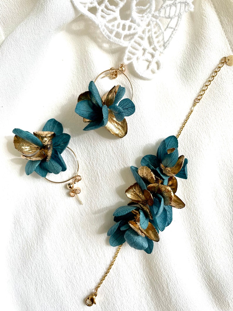 Puede incluir: Un conjunto de pendientes de flores doradas y azul turquesa y una pulsera a juego. Los pendientes son de aro con un racimo de flores sujetas. La pulsera tiene una cadena con un racimo de flores sujetas.