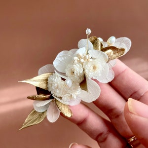 May include: A delicate floral hair comb featuring white dried flowers and gold leaves. The comb is embellished with small pearl accents.