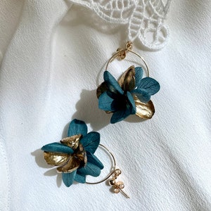 Puede incluir: Un par de pendientes de aro dorados con detalles de flores azules. Las flores est&aacute;n hechas de p&eacute;talos secos y tienen un aspecto delicado y femenino.