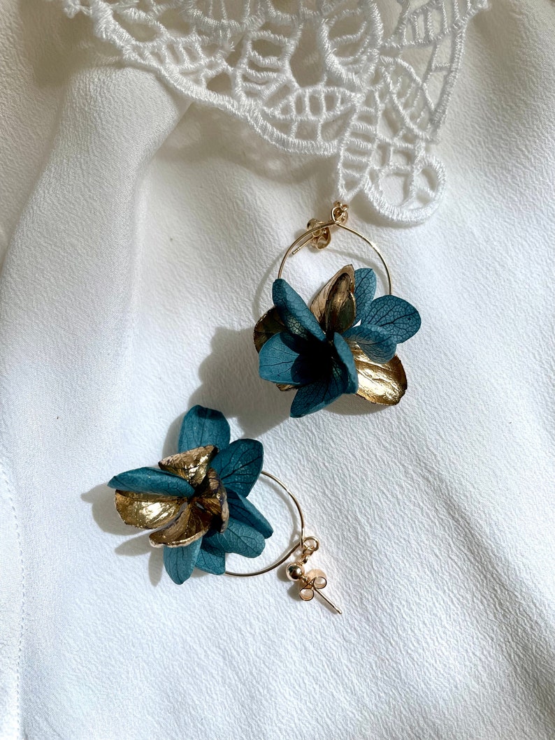 Puede incluir: Un par de pendientes de aro dorados con detalles de flores azules. Las flores est&aacute;n hechas de p&eacute;talos secos y tienen un aspecto delicado y femenino.