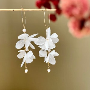 May include: Gold hoop earrings with white flower and pearl details. The earrings have delicate gold chains and small, round pearls, creating an elegant design. The white flowers add a touch of nature-inspired beauty and are suspended from the hoops.