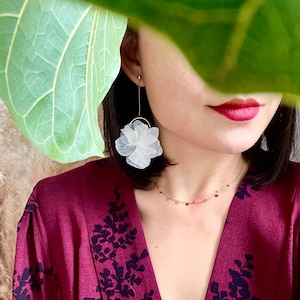 Peut inclure: Gros plan d'une personne portant des boucles d'oreilles blanches en forme de fleur avec des détails dorés. Les boucles d'oreilles sont associées à un collier en or avec de petits charmes circulaires. La personne porte une robe bordeaux avec un motif floral bleu foncé.