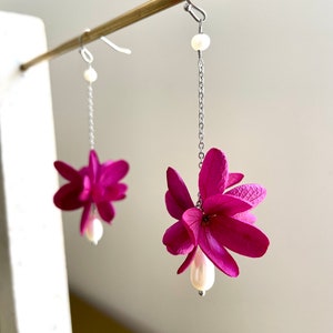 Può includere: Un paio di orecchini pendenti con un gruppo di fiori rosa e una perla bianca in fondo a ogni orecchino.