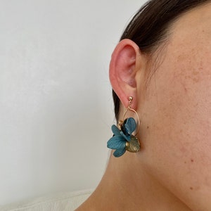 Puede incluir: Un par de pendientes de aro dorados con detalles de flores de tela azul. Los pendientes son colgantes y tienen un aspecto delicado y femenino.