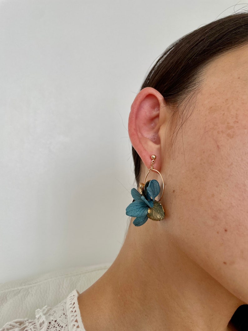 Puede incluir: Un par de pendientes de aro dorados con detalles de flores de tela azul. Los pendientes son colgantes y tienen un aspecto delicado y femenino.