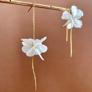 Puede incluir: Un par de pendientes de oro con detalles de flores blancas. Los pendientes cuelgan de una barra de oro.