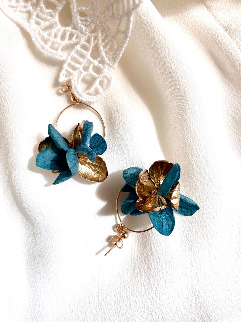 Puede incluir: Un par de pendientes de aro dorados con detalles de flores azules y doradas. Los pendientes est&aacute;n sobre un fondo de tela blanca.