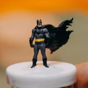 May include: A small, black plastic figurine of Batman standing on a white circular base. The figurine is detailed with a yellow utility belt and a black cape.
