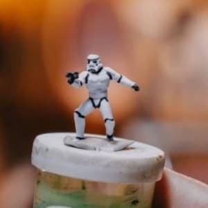 May include: A white plastic figurine of a Stormtrooper from Star Wars, standing on a small, round, grey base. The figurine is holding a blaster in its right hand and has a black visor.