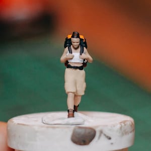 May include: A small figurine of a person wearing a white tank top and khaki shorts, carrying a black and orange backpack. The figurine is standing on a white surface.
