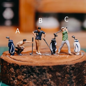 May include: Miniature golf figurines on a wooden base. Three golfers with clubs and bags. The figurines wear blue, green, and tan clothing. The golf bags are black and white or blue and white. The scene is set on a wooden base.