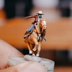 May include: A small figurine depicting a cowboy on horseback. The cowboy is dressed in a white hat, brown jacket, blue shirt, and brown boots. The horse is tan with a white mane and tail. The figurine is mounted on a small, round base.