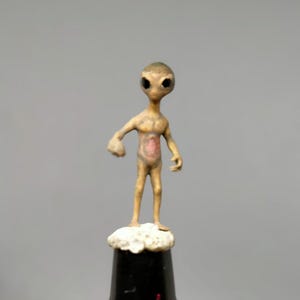 May include: A small, tan-colored alien figurine with large black eyes and a pinkish-red chest. The alien stands on a white cloud-like base atop a black cylindrical object. The figurine has a detailed, textured appearance.