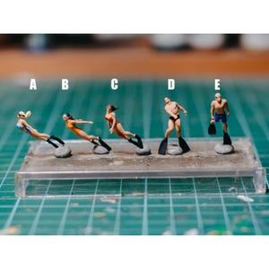 Könnte beinhalten: Miniaturfiguren von Tauchern in Badeanzügen und Flossen. Die Taucher befinden sich in verschiedenen Posen, einige scheinen zu schwimmen. Die Figuren sind auf kleinen, grauen Sockeln montiert und in einer Reihe angeordnet. Der Hintergrund ist ein grünes Gitter.