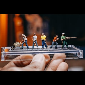 Puede incluir: Cinco figuras en miniatura de personas en varias poses, incluyendo una persona empujando una carretilla, una persona barriendo y una persona sosteniendo una cuerda. Las figuras están pintadas en colores brillantes y se muestran en una base de plástico transparente.
