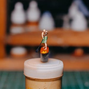 May include: A small figurine of a person paddleboarding on an orange surfboard with a black paddle. The person wears green shorts and stands on a grey cylinder. The background is blurred, with a white lid in the foreground.
