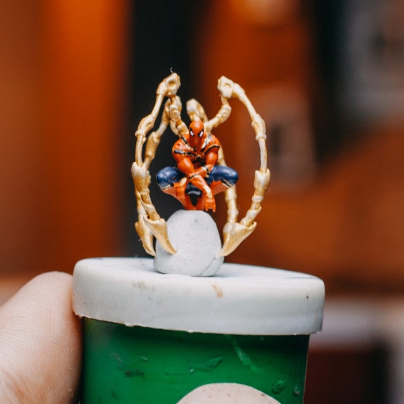 May include: A small, red and blue superhero figurine with gold arms, sitting on a white base. The figurine is on top of a green container with a white lid.