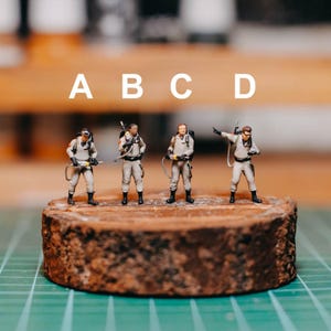 May include: Four miniature figures of Ghostbusters characters standing on a brown wooden platform. The figures are wearing tan jumpsuits and holding proton packs. The figures are labelled A, B, C, and D.