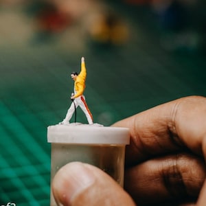 May include: A miniature figurine of a person wearing a yellow jacket and white trousers, holding a microphone and standing on a white platform. The figurine is on a white container.