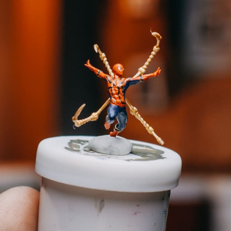 May include: A small, red and blue action figure of Spider-Man with gold web shooters, standing on a small, gray rock. The figure is posed with arms outstretched and legs bent, as if in mid-air. The figure is on top of a white container.