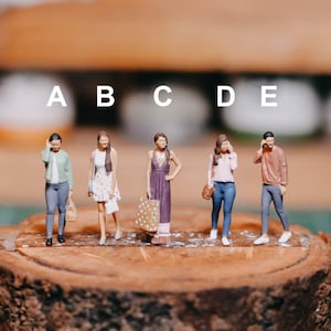 May include: Five miniature figurines of people standing on a wooden log. The figures are wearing different outfits, including a green jacket, a white dress with polka dots, a purple dress, a pink shirt and blue jeans, and a brown shirt and blue jeans. The figures are labelled A through E.