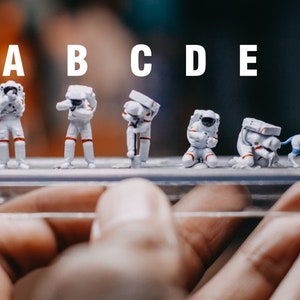 Puede incluir: Figuras de astronautas en miniatura con trajes espaciales blancos y detalles rojos, en varias poses, incluyendo una con un perro. Las figuras se exhiben sobre una base de plástico transparente, sostenida por una mano.