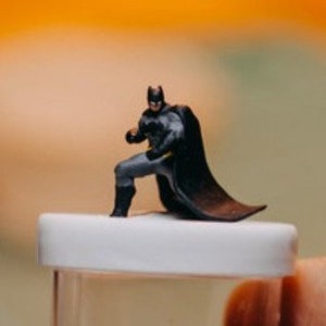 May include: A small, black and grey plastic figurine of Batman in a crouching pose. The figurine is about 5 cm tall and has a cape that is flowing behind him.