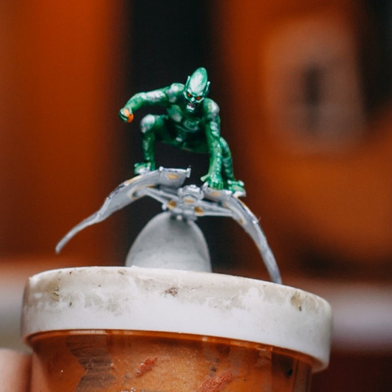 May include: A green and silver action figure of a superhero perched on a silver glider. The figure is posed in a dynamic action stance with its arms outstretched. The figure is perched on a small, silver, and white base.