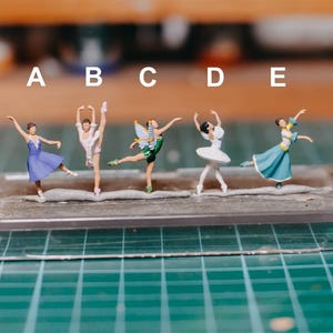 Puede incluir: Figuras en miniatura de bailarinas en varias poses y vestuarios. Las bailarinas visten tonos de azul, blanco y verde, una de ellas adornada con alas de hada. Están dispuestas sobre una base rectangular transparente, sobre una cuadrícula verde.