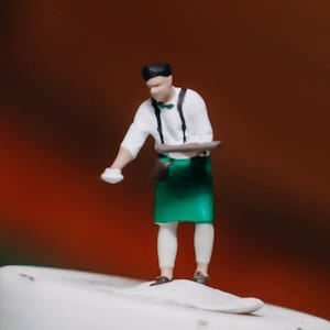 May include: A miniature figurine of a person wearing a white shirt with a black bow tie and a green apron. The person is holding a brown tray in their right hand and a white object in their left hand. The figurine is standing on a white surface.