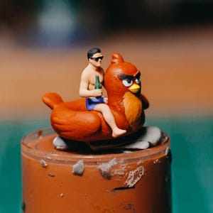 May include: A miniature figurine depicts a person in sunglasses and blue shorts riding a red bird with an angry expression. The person holds a green bottle. The figurine is set on a brown surface.
