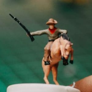 May include: A miniature plastic cowboy riding a rearing horse, both painted in brown and tan tones. The cowboy wears a brown hat, a red bandana, and holds a black rifle.
