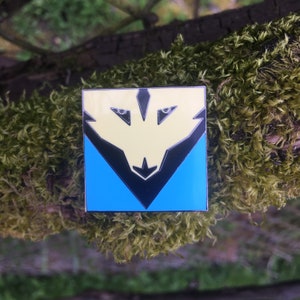 May include: A yellow and blue enamel pin with a black wolf design. The wolf's eyes are white and the pin is square shaped.