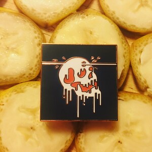 May include: A black enamel pin with a white skull dripping with white and red paint. The skull is set against a dark blue background.