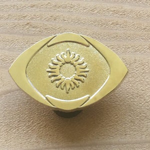 May include: A gold-colored cabinet knob with an oval shape and a sunburst design in the center. The knob is made of metal and has a smooth, polished finish. The knob is mounted on a light brown wooden surface.