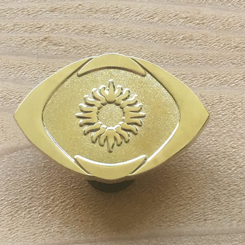 May include: A gold-colored cabinet knob with an oval shape and a sunburst design in the center. The knob is made of metal and has a smooth, polished finish. The knob is mounted on a light brown wooden surface.