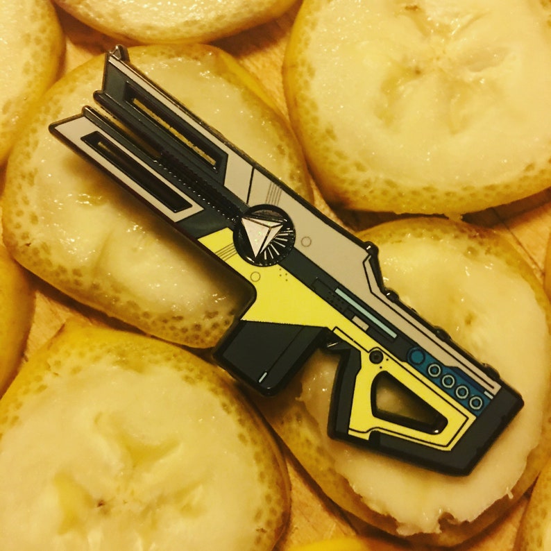 May include: A black, gray, and yellow enamel pin of a futuristic gun with a triangle symbol on the side.