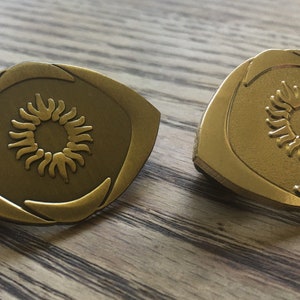 May include: Two bronze-colored, eye-shaped pins with a sunburst design in the center. The pins have a raised border and are set against a wooden surface. The sunburst design is a detailed, stylized representation of the sun.