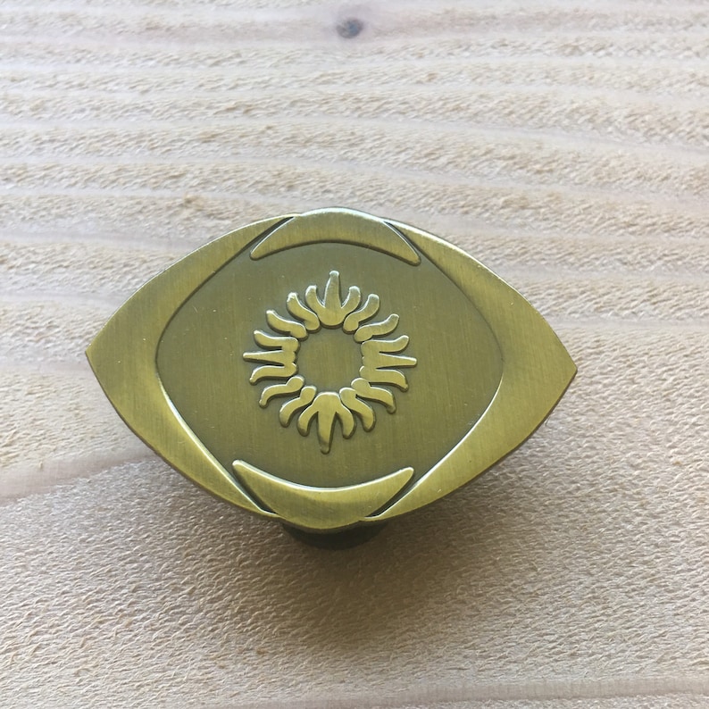 May include: A brass-toned cabinet knob with an eye-shaped design. The center features a sunburst pattern. The knob is mounted on a light-colored wooden surface.