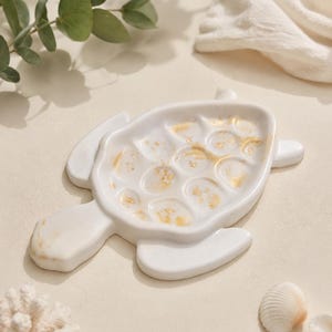 May include: A white turtle-shaped trinket dish featuring a smooth finish with gold leaf accents embedded within the shell section. The dish has a defined head, four flippers, and a textured shell pattern that creates small, shallow circular indentations.