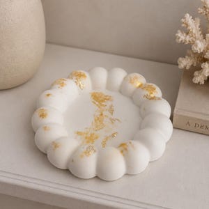 May include: A circular white tray with a scalloped, bubble-like edge and a smooth centre. The surface is decorated with scattered, irregular gold leaf flakes that provide a metallic contrast against the matte white material of the tray.