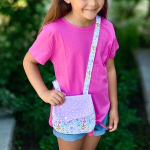 May include: A bright pink short-sleeved t-shirt paired with blue denim shorts and a white headband. A small crossbody bag features a light purple flap with white dots and a lower section with a colorful floral and leaf pattern on a white background.