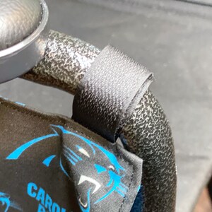 May include: Black fabric with blue Carolina Panthers logos and text is attached to a black metal frame with a black strap. The fabric is wrapped around the frame. The image is a close-up.