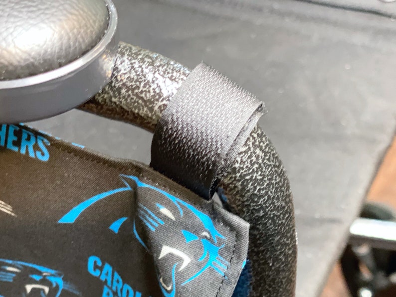 May include: Black fabric with blue Carolina Panthers logos and text is attached to a black metal frame with a black strap. The fabric is wrapped around the frame. The image is a close-up.